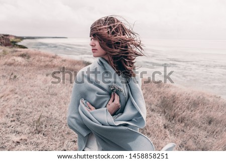 Attractive young woman standing on a windy cold beach wrapped in warm blanket with flower in her hand.