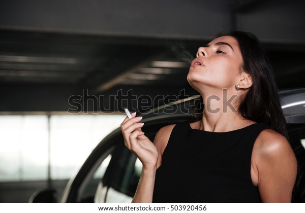 Attractive young woman standing and smoking on\
car parking