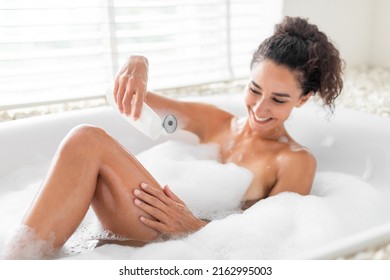 Attractive Young Woman Taking Bath With Foam, Applying Soap On Her Body At Home, Selective Focus. Smiling Millennial Female Pampering Herself, Relaxing In Bathtub, Doing Domestic Beauty Procedure