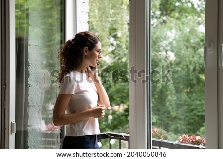 Attractive young woman talk to friend on cellphone while standing near open window at modern home, summer warm day green trees outside. Remote communication using mobile connection, lifestyle concept