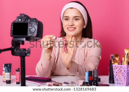 Attractive young woman tests some beauty products and shows lip gloss in her blog. Beauty vlogger sits in front of camera and hods lipstick in her hands . Online translation of tutorial video.