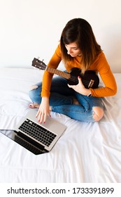 Attractive Young Woman Typing On Her Laptop While Taking Online Ukelele Classes Sitting On Her Bed With A Yellow T-shirt And Jeans.