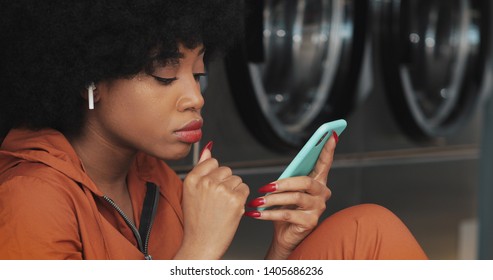 Attractive young woman using smartphone at laundromat. She is wearing a wireless earphones and singing indoors. - Powered by Shutterstock