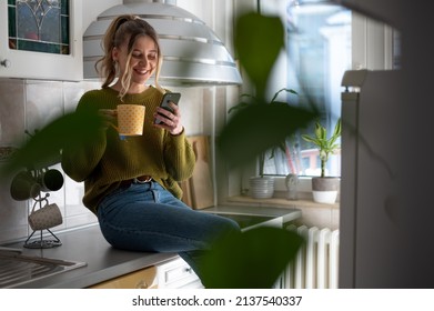 Attractive young woman using a smartphone and drinking cup of tea while standing at the kitchen at home. Concept of leisure activity at home. - Powered by Shutterstock