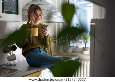 Attractive young woman using a smartphone and drinking cup of tea while standing at the kitchen at home. Concept of leisure activity at home.