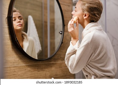 Attractive young woman wearing bathrobe applying makeup with sponge while looking in the mirror in bathroom