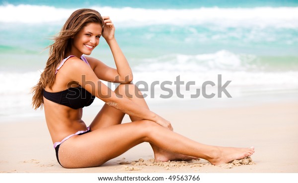 黒いビキニを着た魅力的な若い女性が 膝に肘を乗せ 頭に手を当ててビーチに座る 背景にサーフが見えます 水平ショット の写真素材 今すぐ編集