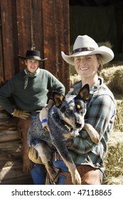 Attractive Young Woman Wearing A Cowboy Hat And Holding An Australian Shepherd. A Young Man Is Standing In The Background. Vertical Shot.