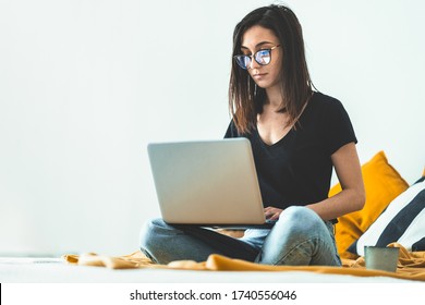 Attractive young woman wearing eye glasses working from home. Female entrepreneur sitting on bed and working from laptop computer while drinking morning coffee. Student learning for exam.