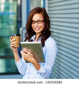 Attractive Young Woman Wearing Glasses Drinking Coffee And Reading Her Touchscreen Tablet While Standing Outside A Commercial Building