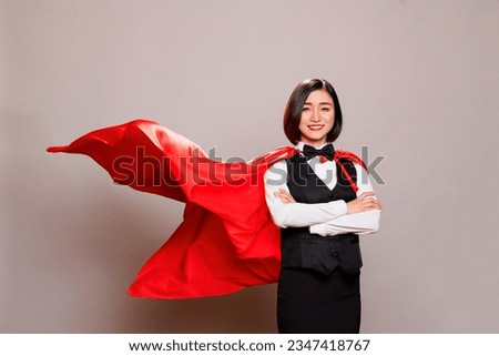 Attractive young woman wearing waitress uniform and fluttering superman cloak while posing in studio portrait. Confident restaurant employee dressed in superhero red cape