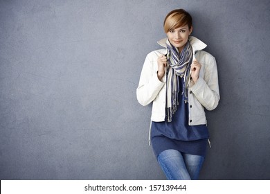 Attractive young woman in white jacket and scarf standing by grey wall