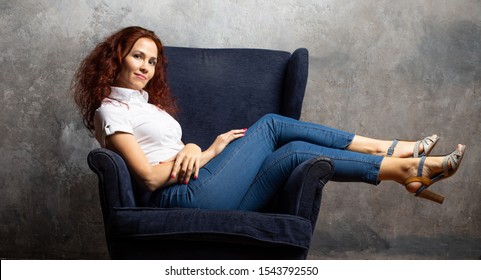 Attractive Young Woman In A White Shirt And Blue Jeans Half Reclining On A Chair On Textured Background