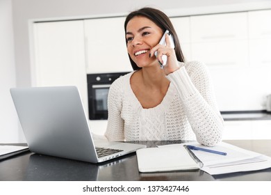Attraktive junge Frau, die mit Laptop-Computer und Dokumenten arbeitet, während sie in der Küche sitzt, mit Mobiltelefon