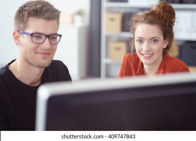 Attractive Young Woman Working In The Office With Her Male Business Partner Sitting At The Desk Looking Over The Computer Monitor At The Camera