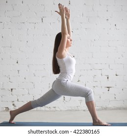 Attractive Young Woman Working Out Indoors. Beautiful Model Doing Exercises On Blue Mat In Room With White Walls. Warrior 1 Or Virabhadrasana I Pose. Square Image. Full Length