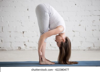 Attractive young woman working out indoors. Side view portrait of beautiful model doing yoga exercise on blue mat. Standing in Uttanasana with palms at the backs of ankles. Full length