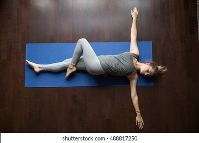 Attractive young woman working out indoors, doing yoga exercise on wooden floor, lying in Reclining Spinal Twist, Jathara Parivartanasana, resting after practice, full length, top view