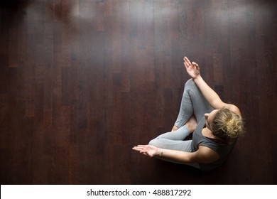 Attractive young woman working out indoors, doing yoga exercise on wooden floor, sitting in Easy (Decent, Pleasant Posture), meditating, breathing, relaxing. Full length. Top view. Copy space - Powered by Shutterstock