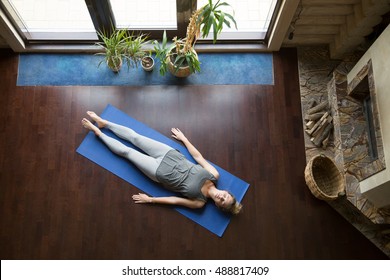 Attractive Young Woman Working Out In Living Room, Doing Yoga Exercise On Blue Mat, Lying In Shavasana (Corpse, Dead Body Posture), Resting After Practice, Meditating, Breathing. Full Length, Top View