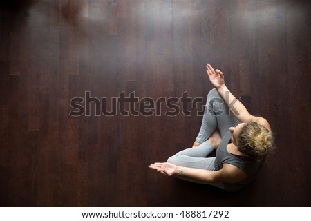Attractive young woman working out indoors, doing yoga exercise on wooden floor, sitting in Easy (Decent, Pleasant Posture), meditating, breathing, relaxing. Full length. Top view. Copy space