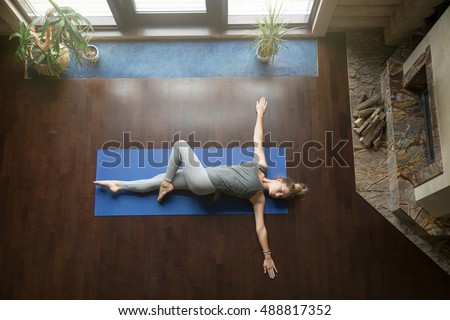 Attractive young woman working out in living room, doing yoga exercise on wooden floor, lying in Reclining Spinal Twist, Jathara Parivartanasana, resting after practice, full length, top view