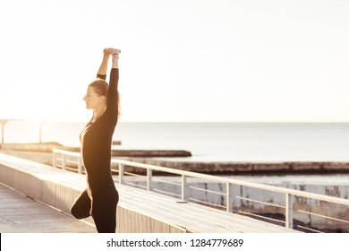 Attractive young yogi woman working out outdoors, Standing in Vrksasana, Tree pose, on open wooden terrace, meditating, breathing, relaxing by the sea on sunrise. Calm and harmony. Morning work-out - Powered by Shutterstock