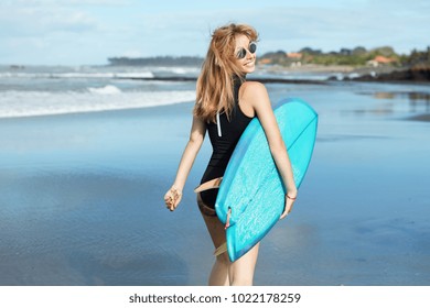 Attrative slender young female model with blonde hair, dressed in swimsuit and wears sunglasses, holds kite board under arm, waits for good conditions on ocean for surfing, walks at coastline - Powered by Shutterstock