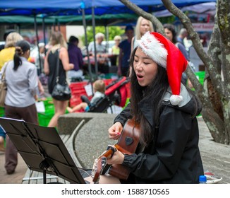 Auckland, New Zealand, December 12 2015. An Attractive Young Asian Woman Wearing A Christmas Hat And Playing A Guitar And Busking At A Farmers Market