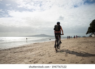 Auckland, New Zealand - February 8 2021: A Man Cycling On The Milford Beach With Rangitoto Island In The Distance. Out Of Focus Background People And Dogs Walking On The Beach.  