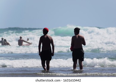 Auckland  New Zealand - January 31 2020: View Of Two Young Polynesian Men In Swimwear Walking In The Water Towards People Playing In Surf At Piha Beach 