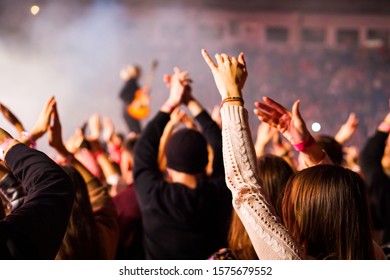 Audience with hands raised at a music festival and lights streaming down from above the stage. Soft focus, high ISO, grainy image. - Shutterstock ID 1575679552