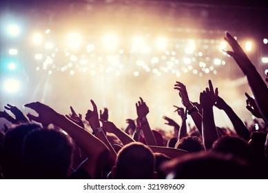 Audience with hands raised at a music festival and lights streaming down from above the stage. Soft focus, high ISO, grainy image.