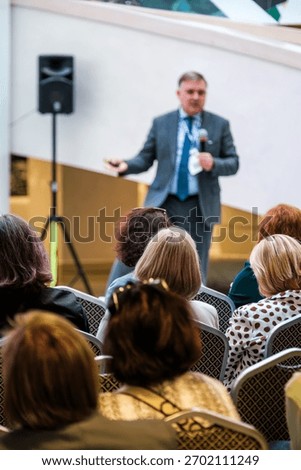 Audience listens while professional speaker presents in conference hall with microphone and presentation setup. Formal attire, attentive crowd, corporate event, business talk, networking opportunities