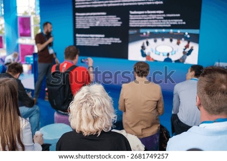Audience listens while speaker presents on large screen in bright blue venue, slides shown, conference vibe, learning, teamwork, tech focus, professional atmosphere
