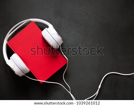 Audio book concept. White headphones and a red book on a stone black desk. Flat lay, top view, copy space