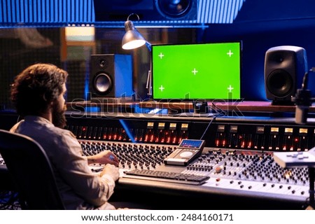 Audio engineer working in studio control room with greenscreen mockup, twisting knobs and switchers to produce music. Producer composer editing tracks and adding sound effects.