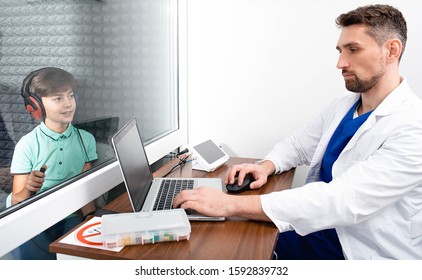 Audiologist Doing Hearing Test To A Little Boy. Child Sitting At Soundproof Cabin, Boy Wearing Headphones Pushing Button While Audiometry Test