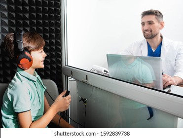 Audiologist Doing Hearing Test To A Little Boy. Child Sitting At Soundproof Cabin, Boy Wearing Headphones Pushing Button While Audiometry Test