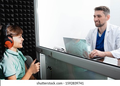 Audiologist Doing Hearing Test To A Little Boy. Child Sitting At Soundproof Cabin, Boy Wearing Headphones Pushing Button While Audiometry Test