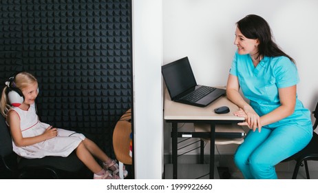 Audiologist Doing A Hearing Test For A Little Girl. Child Sitting At Soundproof Cabin Wearing Headphones Pushing Button While Audiometry Test