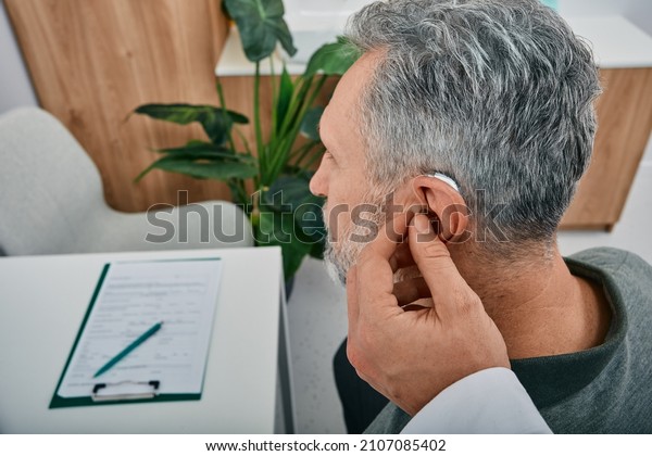 Audiologist fits a\
hearing aid on deafness mature man ear while visit a hearing\
clinic. Hearing solutions for\
older