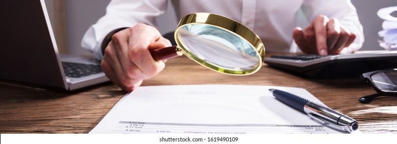 Auditor Analyzing Bill Through Magnifying Glass On Desk