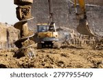 Auger drill bit pulling soil out of a hole in the ground during construction work, with an excavator and a bare brick wall in the soft-focus background
