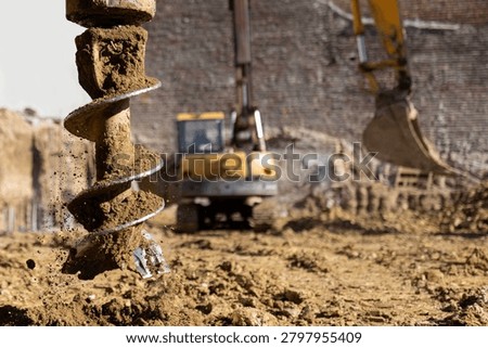 Auger drill bit pulling soil out of a hole in the ground during construction work, with an excavator and a bare brick wall in the soft-focus background