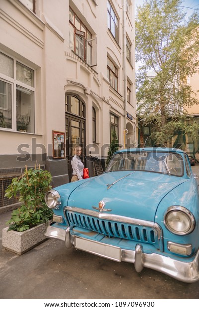 August 19, 2020,\
Stary Arbat, Moscow, Russia. Beautiful young woman standing in the\
yard by a blue retro car