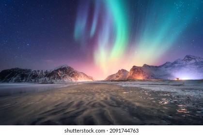 Aurora borealis above the snowy mountain and sandy beach in winter. Northern lights in Lofoten islands, Norway. Starry sky with polar lights. Night landscape with aurora, frozen sea coast, city lights - Powered by Shutterstock