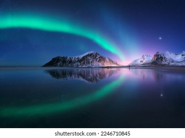 Aurora borealis and beach in Lofoten islands, Norway. Beautiful northern lights. Starry sky with polar lights. Night winter landscape with aurora, sea with sky reflection, city lights, snowy mountains