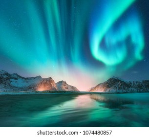 Aurora borealis in Lofoten islands, Norway. Aurora. Green northern lights. Starry sky with polar lights. Night winter landscape with aurora, sea with sky reflection, stones, beach and snowy mountains - Shutterstock ID 1074480857