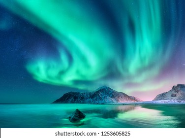 Aurora borealis. Northern lights in Lofoten islands, Norway. Sky with polar lights and stars. Night winter landscape with aurora, sea with sky reflection, stones, sandy beach and snowy mountains - Shutterstock ID 1253605825